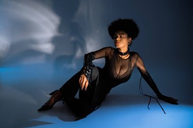 Kıvırcık Afro-Amerikan kadın şeffaf gömlekli ve seksi kıyafetli mavi üzerinde oturuyor. 