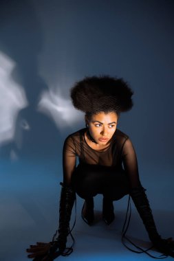 Modayı takip eden Afro-Amerikan kadın şeffaf gömlekli ve seksi kıyafetli, mavi üzerinde otururken başka tarafa bakıyor. 