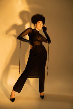 Tam boy, kıvırcık, Afro-Amerikan modelli, seksi, şeffaf kıyafetli, kalçasında sarı bir poz veren. 