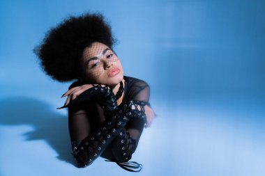 Siyah peçeli genç Afro-Amerikan modeli mavi arka planda poz verirken kameraya bakıyor. 