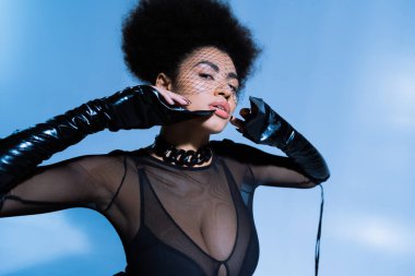 Siyah elbiseli ve peçeli genç Afro-Amerikan kadın mavi poz verirken kameraya bakıyor. 