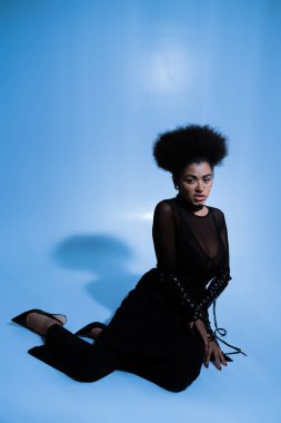 Hoşnutsuz Afro-Amerikan kadın seksi giyinmiş ve siyah peçeli. Mavi üzerine oturmuş kameraya bakıyor. 