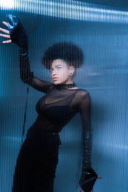 Genç Afro-Amerikalı seksi siyah elbiseli kadın fırfırlı camın arkasında poz veriyor.