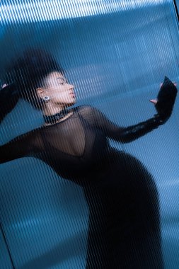 Genç Afro-Amerikan modeli, siyah seksi elbisesiyle dalgalı camların arkasında poz veriyor.