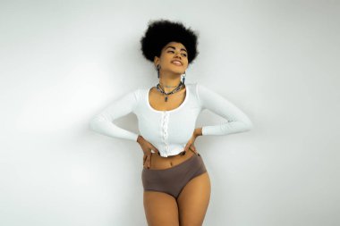 Uzun kollu gömlekli ve iç çamaşırlı mutlu Afro-Amerikan kadın elleri beyaz duvarın yanında duruyor. 