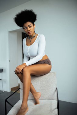 Beyaz duvarın yanındaki koltukta otururken uzun kollu gömlekli ve külotlu genç Afrikalı Amerikalı kadın gözlerini kaçırıyor. 