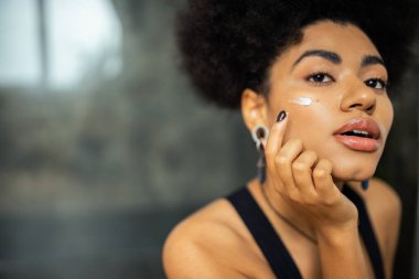 Young african american woman applying cosmetic cream and looking away in bathroom  clipart