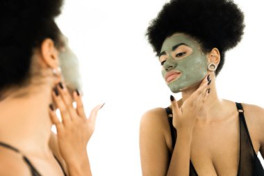 Yüzünde kil maske olan kıvırcık Afrikalı Amerikalı kadın aynaya bakıyor. 