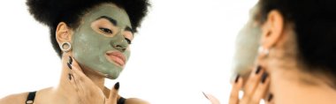 Yüzünde kil maske olan genç Afrikalı Amerikalı kadın banyodaki aynaya bakıyor. 