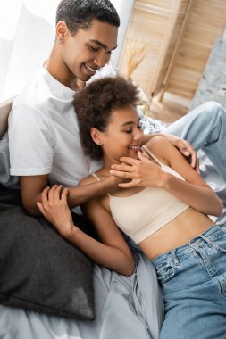 Gülümseyen Afrikalı Amerikalı erkek, seksi kız arkadaşına sarılıyor ve yatakta kot pantolon giyiyor.