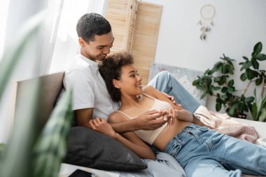 Genç Afro-Amerikalı erkek, seksi kız arkadaşıyla el ele tutuşuyor. Ekin bluzu ve kotuyla yatakta yatıyor.