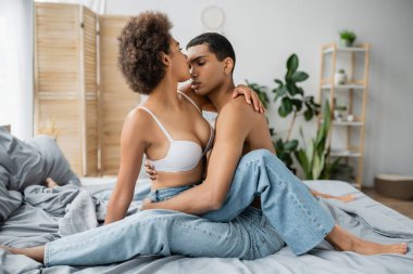 Genç Afro-Amerikan kadın seksi vücutlu, gömleksiz erkek arkadaşı kot pantolonla yatakta oturuyor.