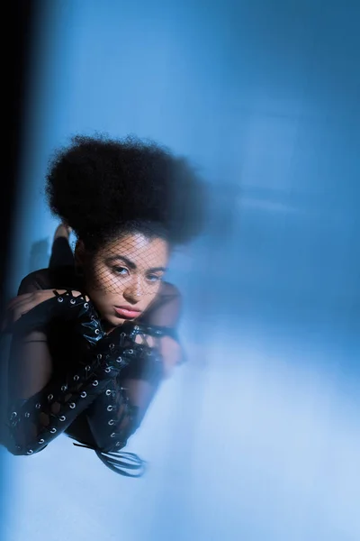 stock image curly african american woman in black veil looking at camera while posing on blue with blurred foreground 