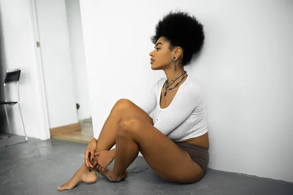 穿着长袖衬衫和内裤坐在白墙附近灰蒙蒙的地板上的非洲裔美国女人的侧视图 — 图库照片