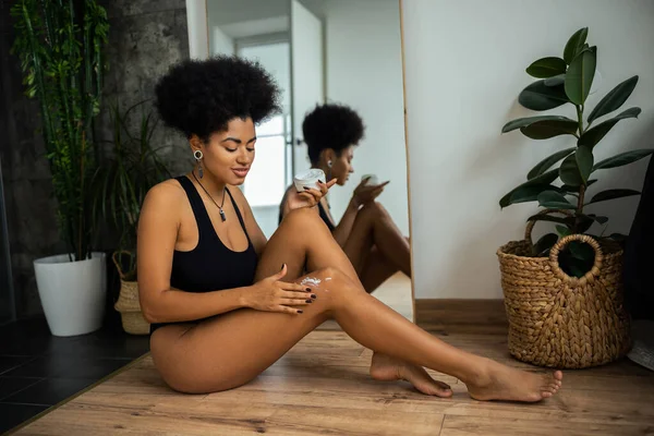 アフリカ系アメリカ人女性が鏡の近くの膝の上に化粧品クリームを適用し バスルームで植物 — ストック写真