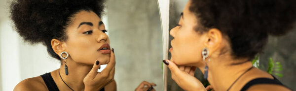Side view of african american woman touching chin while looking at mirror at home, banner 