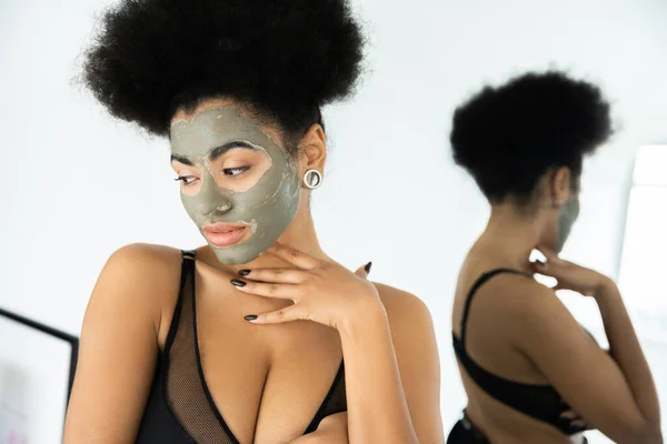 年轻的非洲裔美国女人 脸上戴着黏土面具 站在家里镜子旁边 — 图库照片
