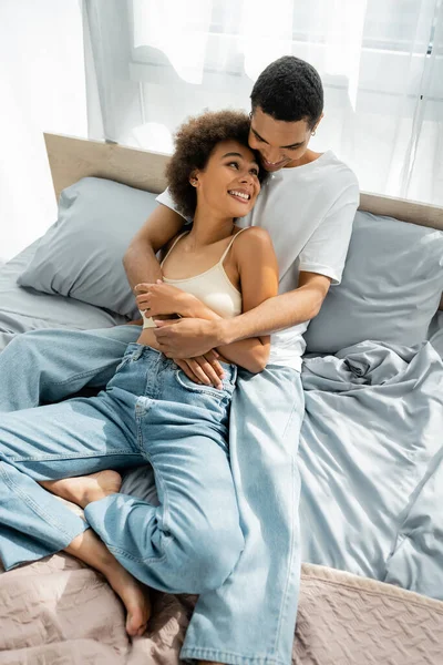 穿着牛仔裤拥抱对方并在床上相互凝视的快乐的非洲裔美国夫妇的高视角照片 — 图库照片