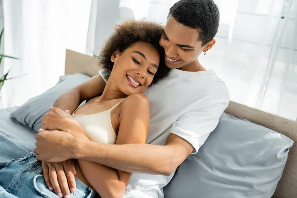 穿着白色T恤衫 抱着快乐的年轻女友躺在床上的非洲裔美国男人 — 图库照片