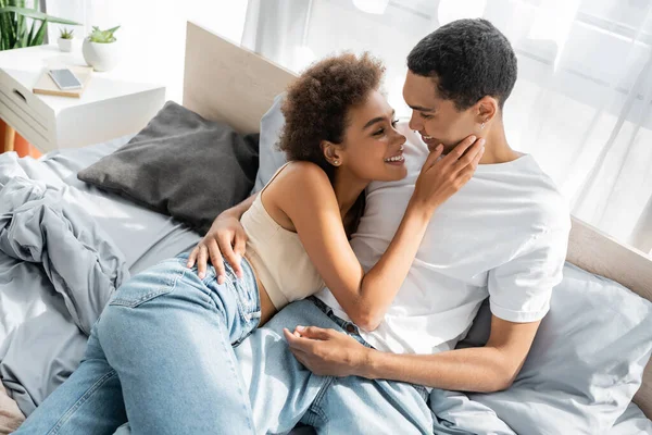 穿着牛仔裤在床上触摸年轻男友脸庞的快乐的非洲女人的高透视 — 图库照片