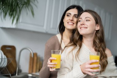 Mutfakta elinde portakal suyu bardakları olan aynı cinsten gülümseyen çift. 