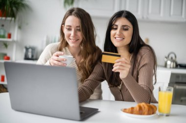Mutfakta kahvaltıda laptopunu ve kredi kartını kullanan gülümseyen kadın. 