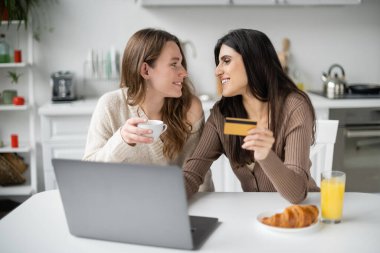 Mutfakta laptopun ve kahvaltının yanında kredi kartı ve kahve tutan aynı cinsten gülümseyen çiftin yan görüntüsü. 