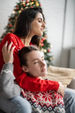 Evde Noel süveteri giymiş kız arkadaşını kucaklayan genç lezbiyen kadın. 