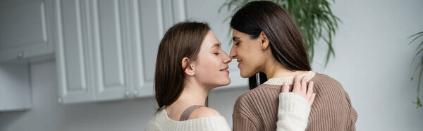 Side view of young same sex couple standing nose to nose in kitchen, banner 