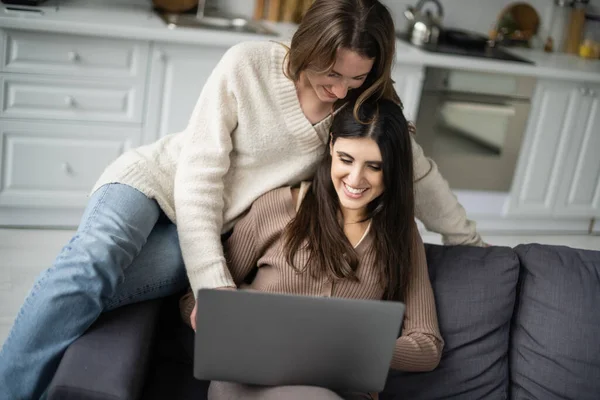 穿着保暖衣服的快乐的女同性恋夫妇在沙发上使用笔记本电脑 — 图库照片