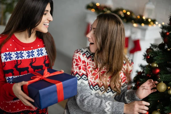 Γυναίκα Πουλόβερ Κρατώντας Χριστουγεννιάτικο Δώρο Κοντά Έκπληκτος Φίλη Στο Σαλόνι — Φωτογραφία Αρχείου