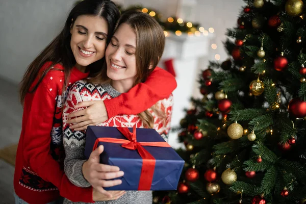 Mujer Positiva Abrazando Novia Con Caja Regalo Durante Celebración Navidad —  Fotos de Stock