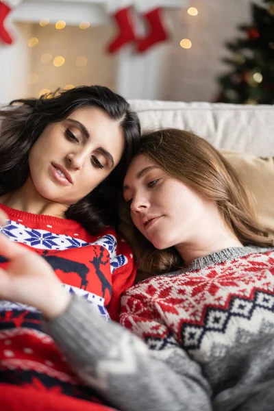 Young lesbian couple in christmas sweaters relaxing on couch at home