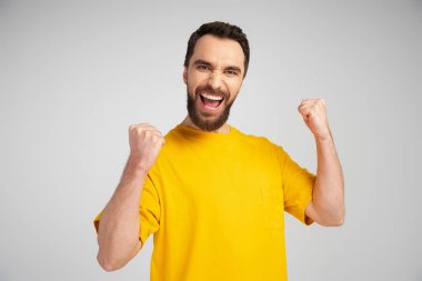 Sarı tişörtlü heyecanlı adam bağırıyor ve gri üzerinde başarı işareti gösteriyor.