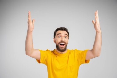 amazed man in yellow t-shirt standing with open mouth and showing wow gesture isolated on grey clipart