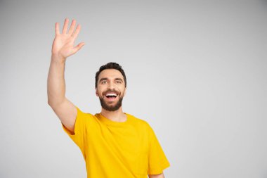 cheerful bearded man in yellow t-shirt waving hand and looking away isolated on grey clipart