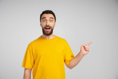 surprised man in yellow t-shirt looking at camera and pointing with finger isolated on grey clipart