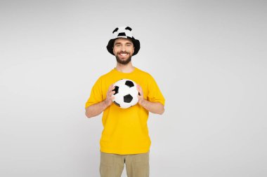 Futbol şapkalı neşeli sakallı adam futbol topu tutuyor ve gri renkli kameraya bakıyor.