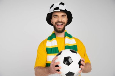 Futbol şapkalı, çizgili eşarplı, topu tutan ve gri renkli kameraya gülen heyecanlı bir adam.