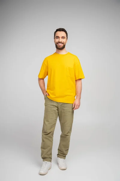 全长的快乐胡子男人 穿着米黄色的裤子和黄色的T恤 手插在口袋里 灰色背景 — 图库照片