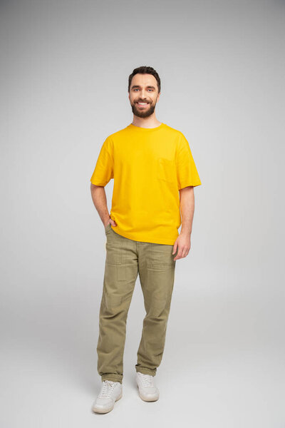 full length of happy bearded man in beige pants and yellow t-shirt standing with hand in pocket on grey background