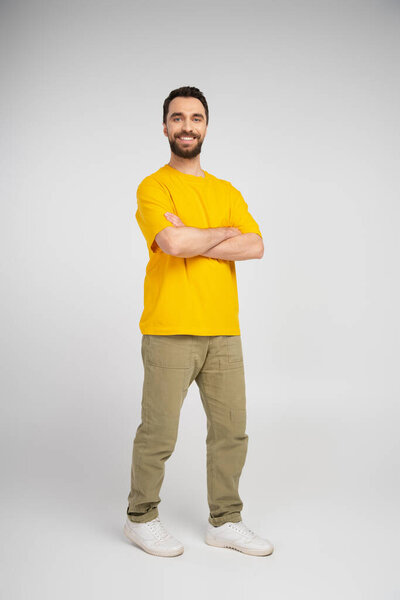 full length of happy bearded man in yellow t-shirt and beige pants standing with crossed arms on grey background