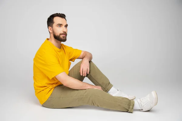 全长的男人穿着米黄色的裤子和黄色的T恤 坐在灰色的背景上看着别处 — 图库照片