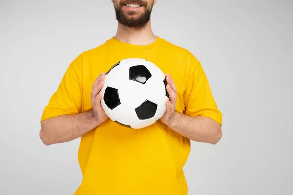 partial view of smiling bearded man in yellow t-shirt holding soccer ball isolated on grey