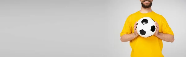 cropped view of bearded and smiling man in yellow t-shirt holding soccer ball isolated on grey, banner