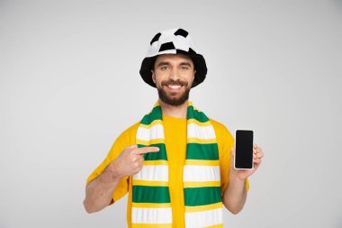 Futbol fanatiği şapkalı mutlu sakallı adam gri ekranlı akıllı telefonu işaret ediyor.
