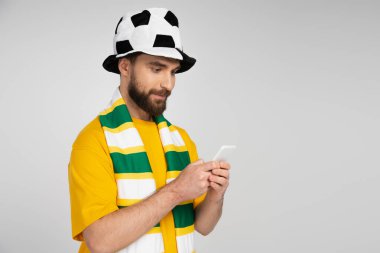 Futbol şapkalı ve çizgili atkılı odaklanmış bir adam gri renkte izole edilmiş akıllı telefondan spor bahisleri oynuyor.