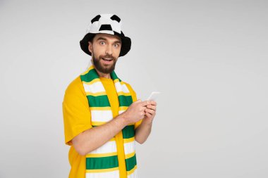 Futbol şapkalı, çizgili fularlı, akıllı telefonlu ve gri renkli kameraya bakan pozitif ve meraklı bir adam.
