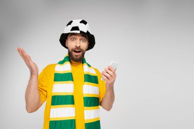 Çizgili eşarplı ve futbol şapkalı bir adamın elinde cep telefonu tutarken gri renkli kameraya bakmasına şaşırdım.