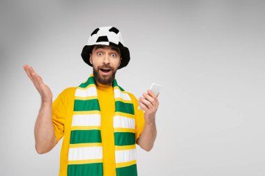 surprised man in striped scarf and football fan hat holding cellphone and looking at camera isolated on grey clipart